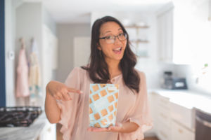 Carrie Abbott holds a bag of Frittle in a kitchen. She is wearing a peach colored top and wears glasses.