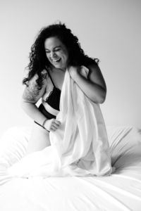 Britnee kneeling on a bed in a bra and underwear, smiling and covering herself with a bedsheet