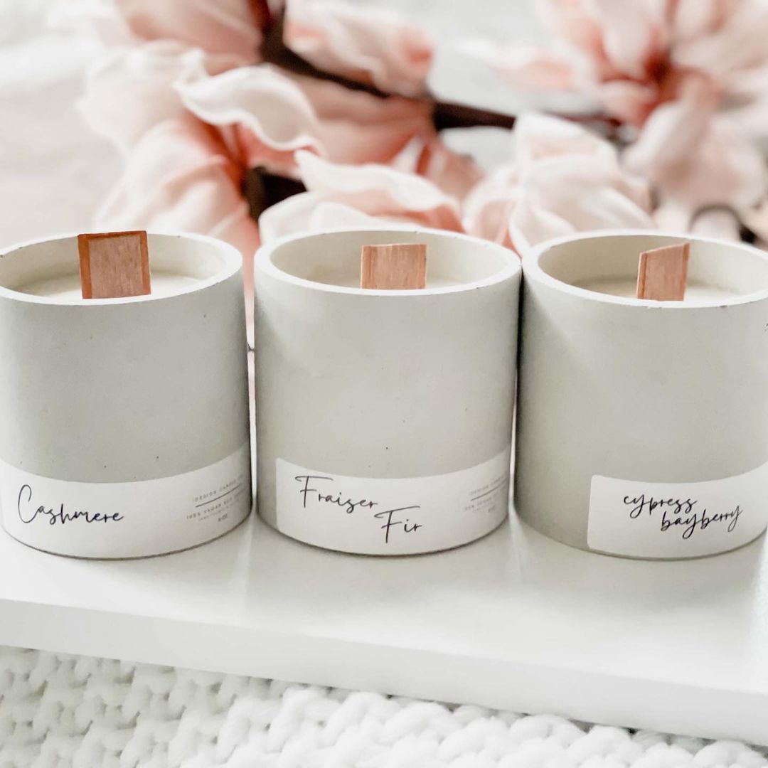 Three light grey candles displayed in a row. Pink flowers in the background.