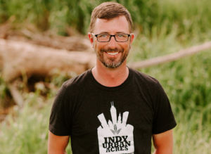 A photo of a man in glasses standing in a field and wearing an Indy Urban Acres t-shirt
