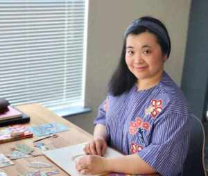 A photo of paper artist Shelley Hanmo Qian sitting at a table with artwork around her