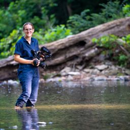 A photo of a woman holding a camera and standing in a creek