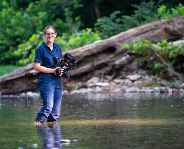 A photo of a woman holding a camera and standing in a creek