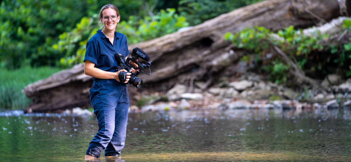 A photo of a woman holding a camera and standing in a creek