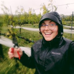 A photo of a woman from Oliver Winery in a rain coat smiling with clippers