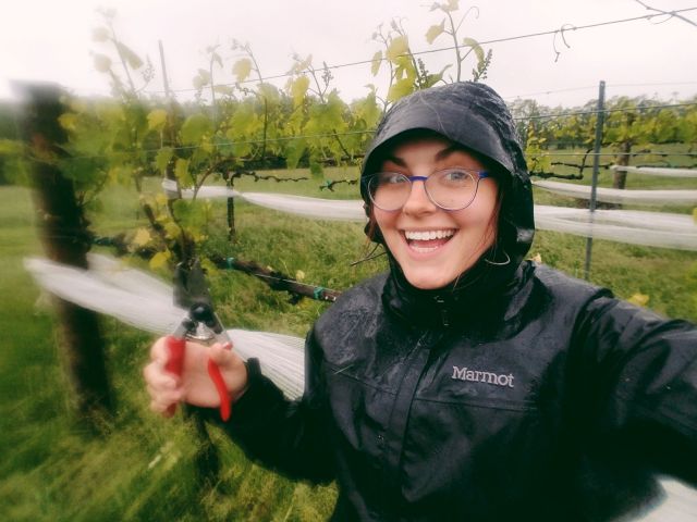 A photo of a woman from Oliver Winery in a rain coat smiling with clippers