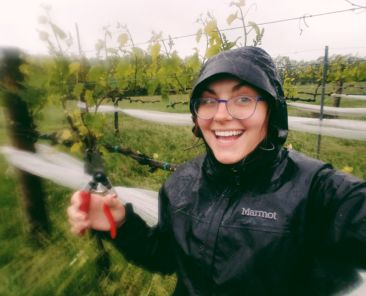A photo of a woman from Oliver Winery in a rain coat smiling with clippers