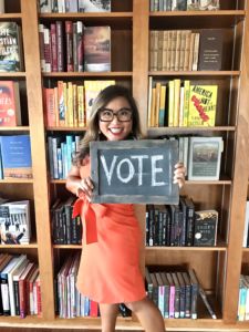 A photo of a woman wearing glasses standing in front of a bookshelf holding a chalkboard that says VOTE