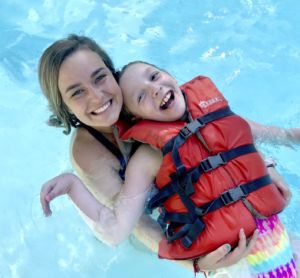 Riley camper wearing a life-jacket having fun swimming in the pool as another camper holds her