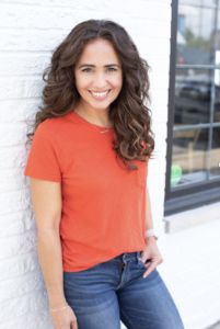 woman with curly brown hair in bright orange shirt stands in front of white brick wall and smiles for the camera