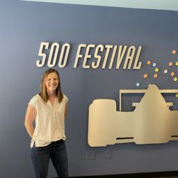 woman wearing white blouse and jeans stands in front of a grey wall with a cutoff of a race car for the indy 500 festival