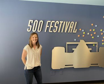 woman wearing white blouse and jeans stands in front of a grey wall with a cutoff of a race car for the indy 500 festival