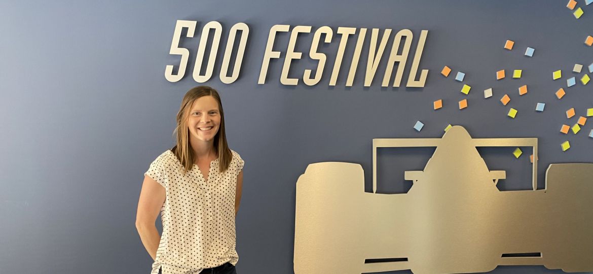 woman wearing white blouse and jeans stands in front of a grey wall with a cutoff of a race car for the indy 500 festival