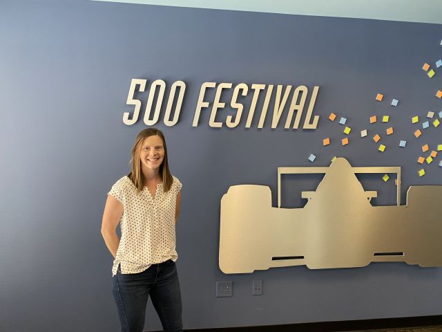 woman wearing white blouse and jeans stands in front of a grey wall with a cutoff of a race car for the indy 500 festival