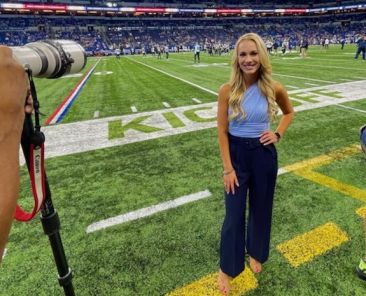 blonde petite woman wearing blue pants and a blue tank top poses with left hand on hip on a football field