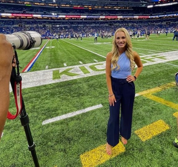 blonde petite woman wearing blue pants and a blue tank top poses with left hand on hip on a football field