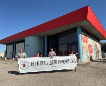 four people hold up a white banner in front of culture center with red roof and artwork on the side of the building