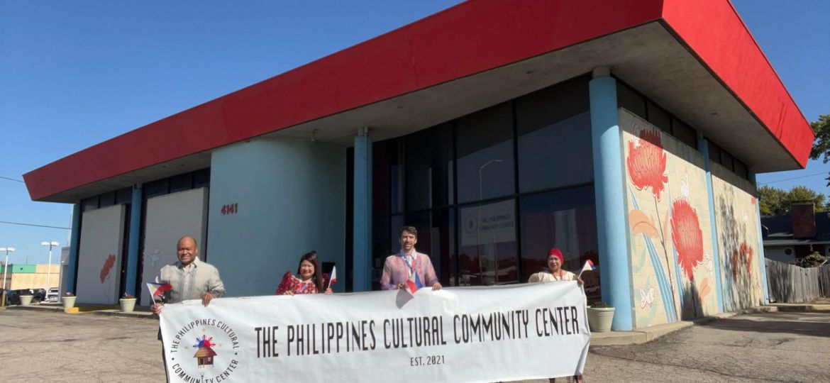 four people hold up a white banner in front of culture center with red roof and artwork on the side of the building