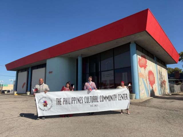 four people hold up a white banner in front of culture center with red roof and artwork on the side of the building