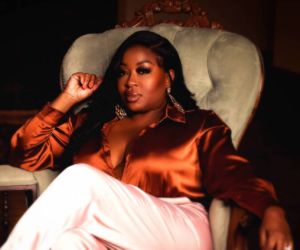 black woman in silk orange collared blouse with white jeans posing mysteriously in a suede chair