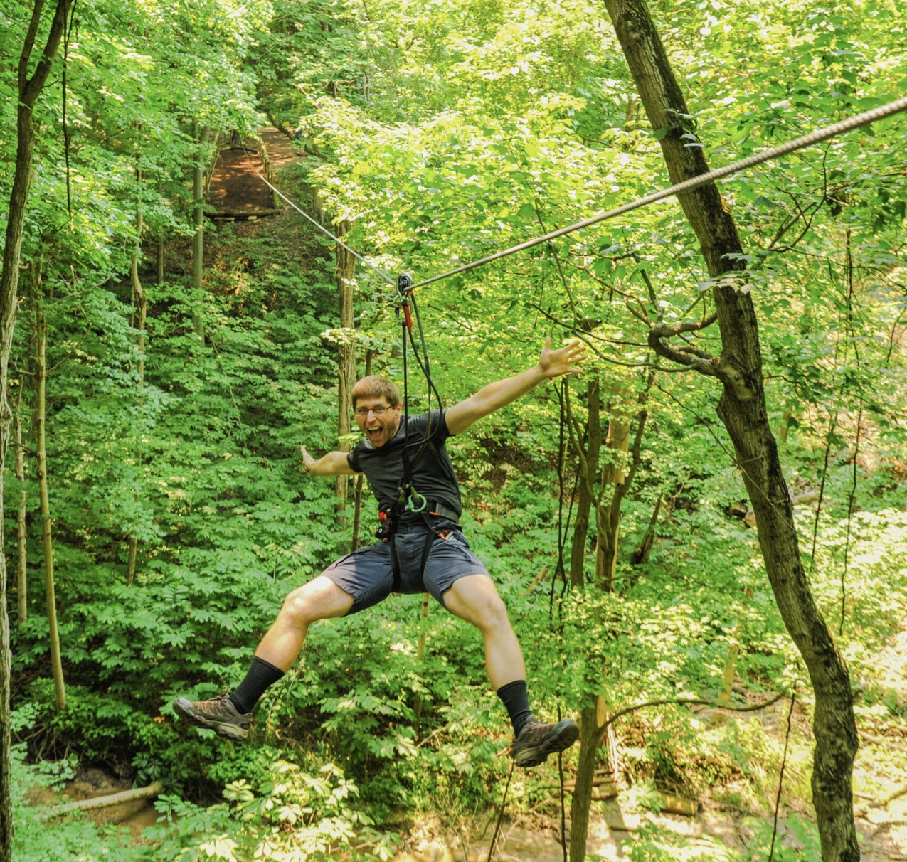 a man in a dark shirt and shorts ziplining through a green forest