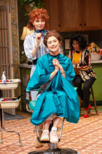 two white women in a play on stage it is set in a hair salon they are both wearing blue