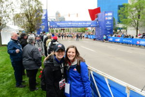 two women stand in front of a blue marathon finish line wearing big winter coats and they are smiling
