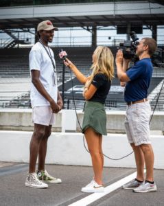 woman in green tennis skirt and black shirt with blonde hairs holds up microphone to interview tall black man wearing white shirt and shorts on live television