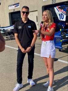 woman with blonde hair is wearing a white skirt and red shirt interviewing a race car driver 