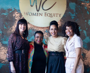 A photo of four women standing in front of a women equity sign smiling