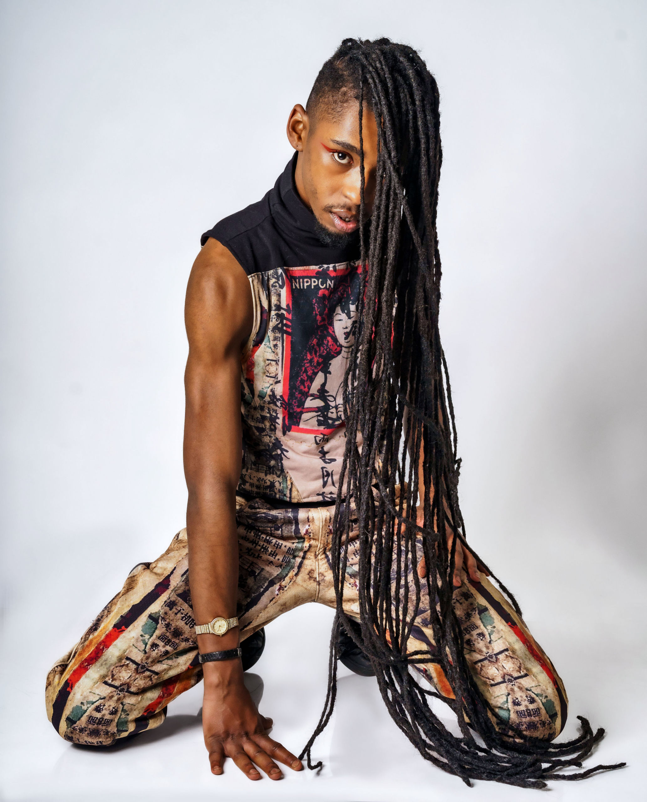 black man with long black dreadlocks poses on knees wearing printed shirt and pants against white background