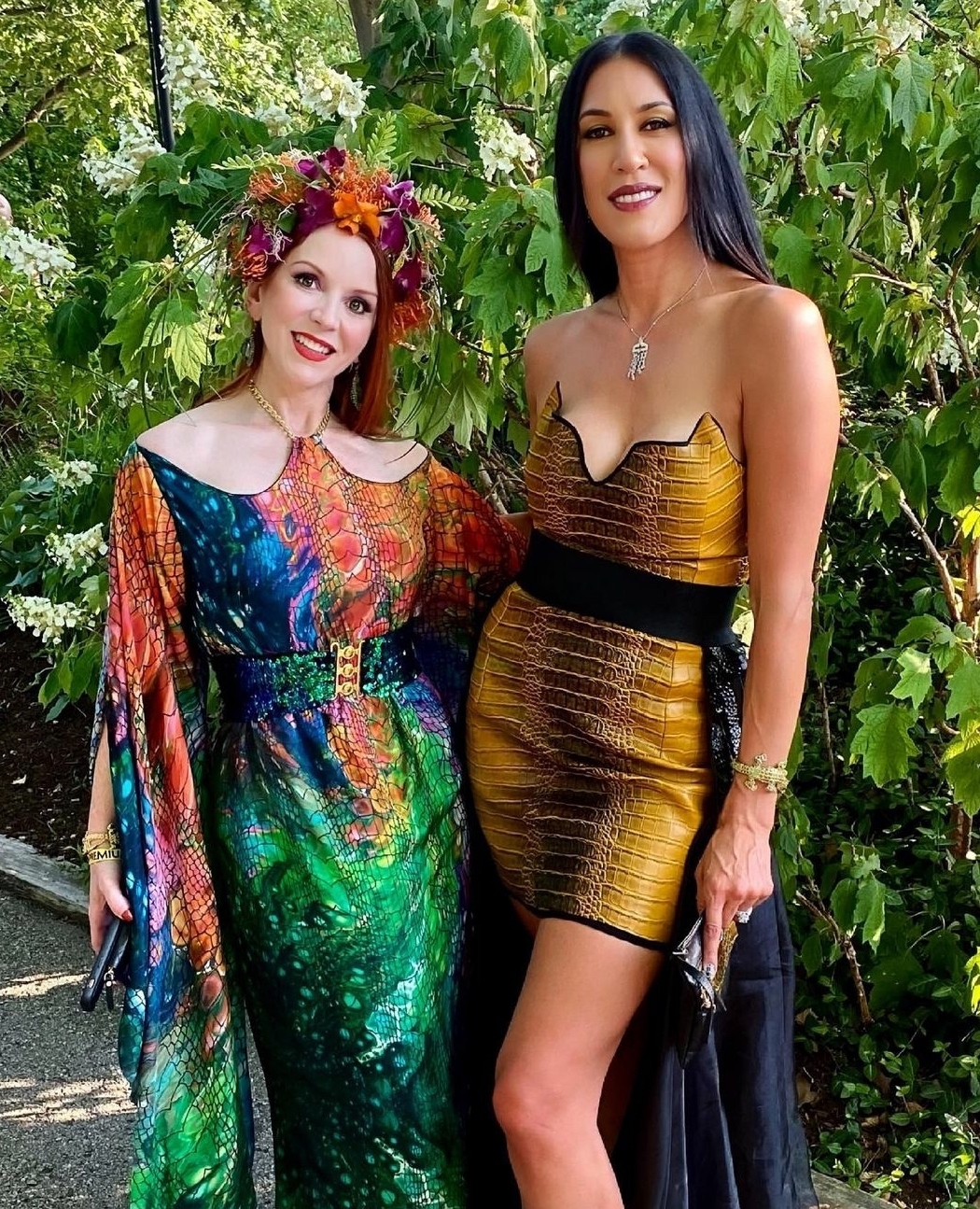 a white woman with red hair wearing a flower crown and a multi-colored dress next to a tall white woman with black hair wearing a golden dress