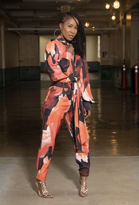 black woman wearing a red orange and brown jumpsuit with gold heels