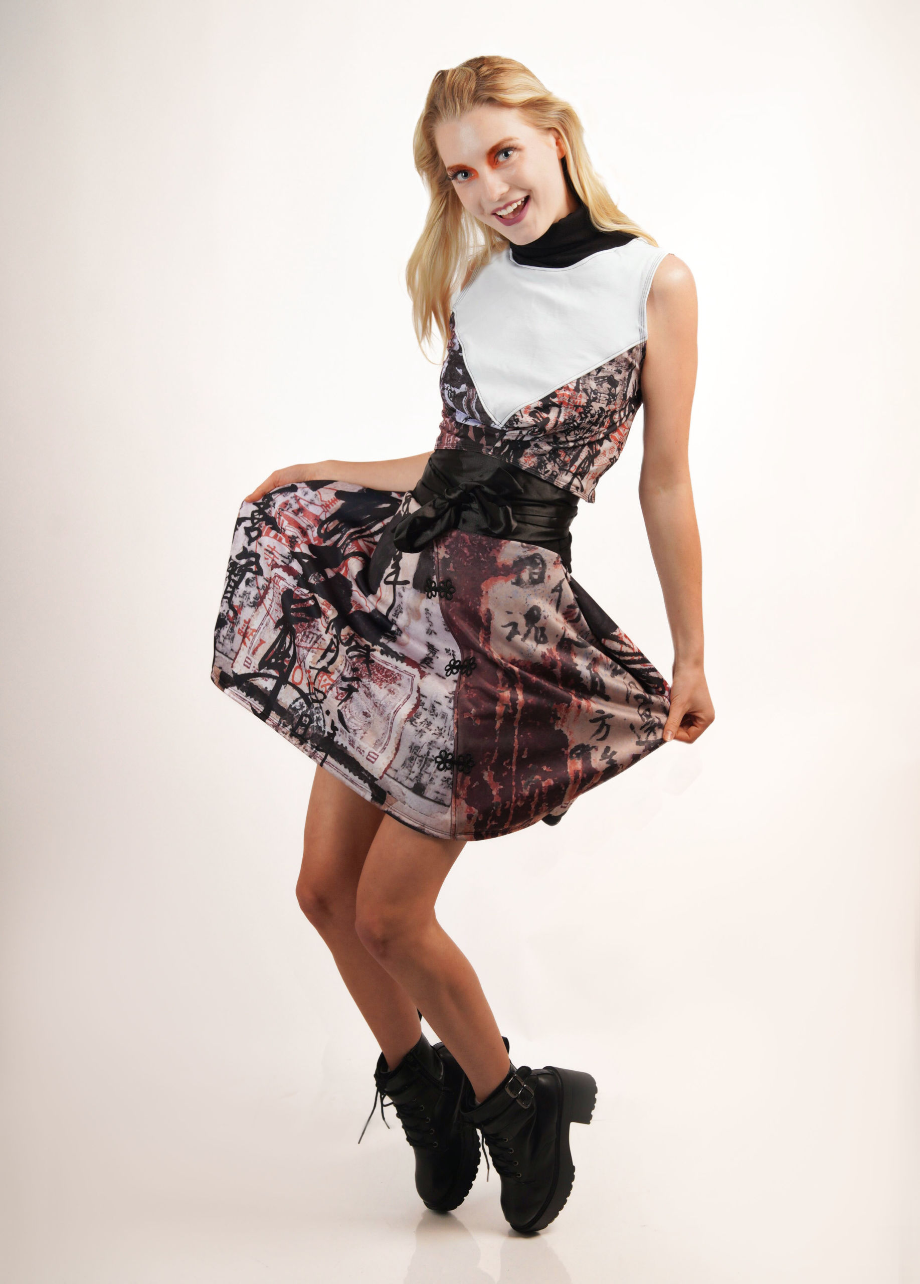 a white woman with medium length blonde hair wearing a multicolored and white printed dress with brown booties posing on her tip-toes