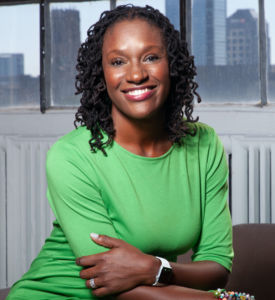 Black woman smiling with teeth wearing a bright green quarter length t-shirt with arms crossed over elbows in front of city buildings