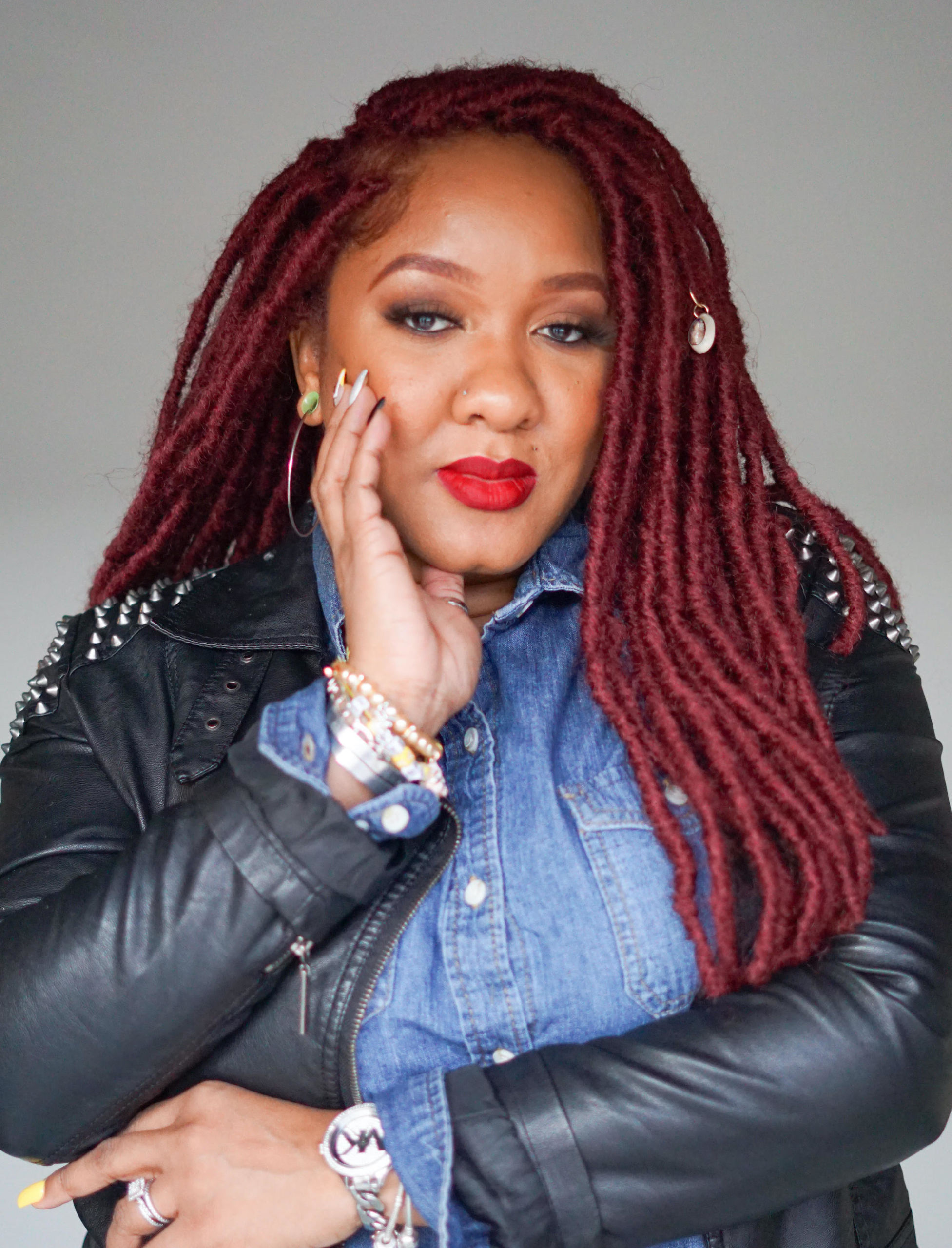 Black woman with long red dreadlocks wears denim shirt with black leather jacket over and looks serious at the camera