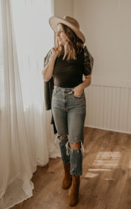 white woman standing by a window with a white curtain wearing a straw hat and ripped jeans and a black t-shirt with a blazer thrown over her shoulder