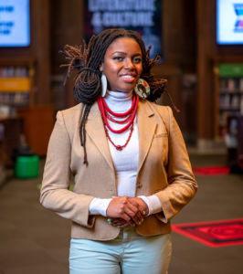 black woman wearing a tan blazer and white turtle neck with a big red necklace posing smiling with teeth holding her hands together in front of her stomach