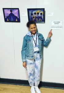 black woman with hair in bun wears jean jacket and joggers pointing next to a sign in an art museum 