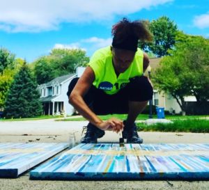 black woman wearing a bright yellow t-shirt bends down outside while painting on a canvas