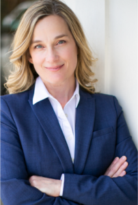white woman with short blonde hair soft smiling for the camera wearing a navy blue blazer with a white collared shirt underneath with arms crossed across her chest