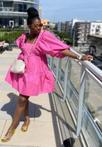 black woman posing on top of an apartment complex wearing a hot pink flowy babydoll dress with yellow sandals and a white cross over body bag white black dreads tied up in a high bun