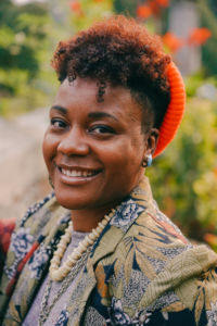 black woman smiling with teeth with a pixie cut wearing an orange beanie and a floral blazer