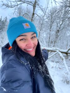 woman with black hair smiling with teeth with blue carhart hat in the woods with snow