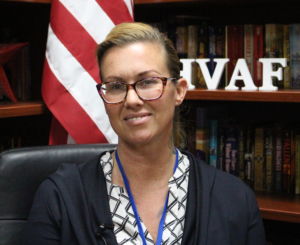 white woman wearing a zig zag patterned blouse, black jackets, wearing glasses smiling with teeth with an American flag behind her 