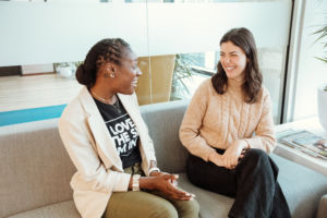 Two women sitting on a couch talking. The one on the left is wearing a blazer and a black t-shirt with green pants. She's smiling at a woman to the right wearing a cream cable knit sweater and black pants smiling back at her.