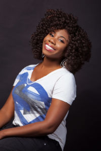black woman with thick short curly brown hair wearing a white t-shirt with a blue design on it smiling with teeth and leaning back from the camera