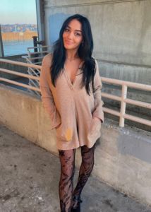 woman with black hair wearing beige romper with black lace tights with hands in her pockets
