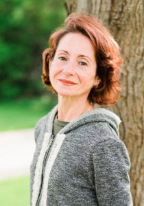 white petite woman wearing a grey sweater and soft smiling in front of a tree trunk she has short red hair