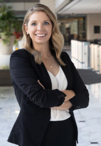 white woman white medium length blonde hair smiling with teeth wearing a matching black blazer and pants set with a white blouse underneath with arms folded across her chest